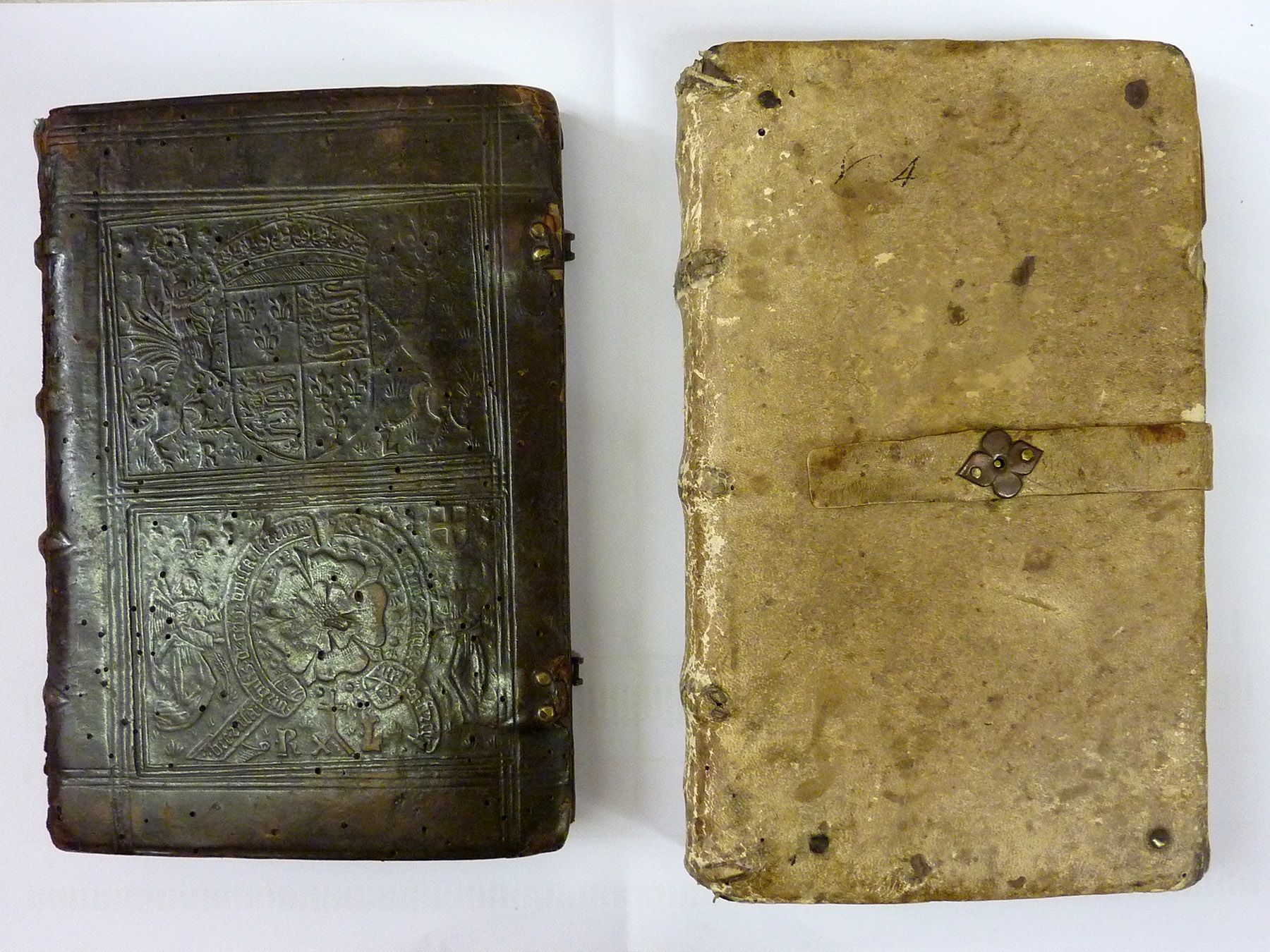Photograph of two very old books side by side. Both are bound in leather. The book on the left is a little smaller and bound in a very dark brown leather which has a complicated design embossed onto it, with a Tudor royal crest and Tudor rose. The book on the right is a lighter creamy colour and much simpler. Its only decoration is a strap to hold the cover close which has a small metal ornament on it, shaped a bit like a flower with four petals.