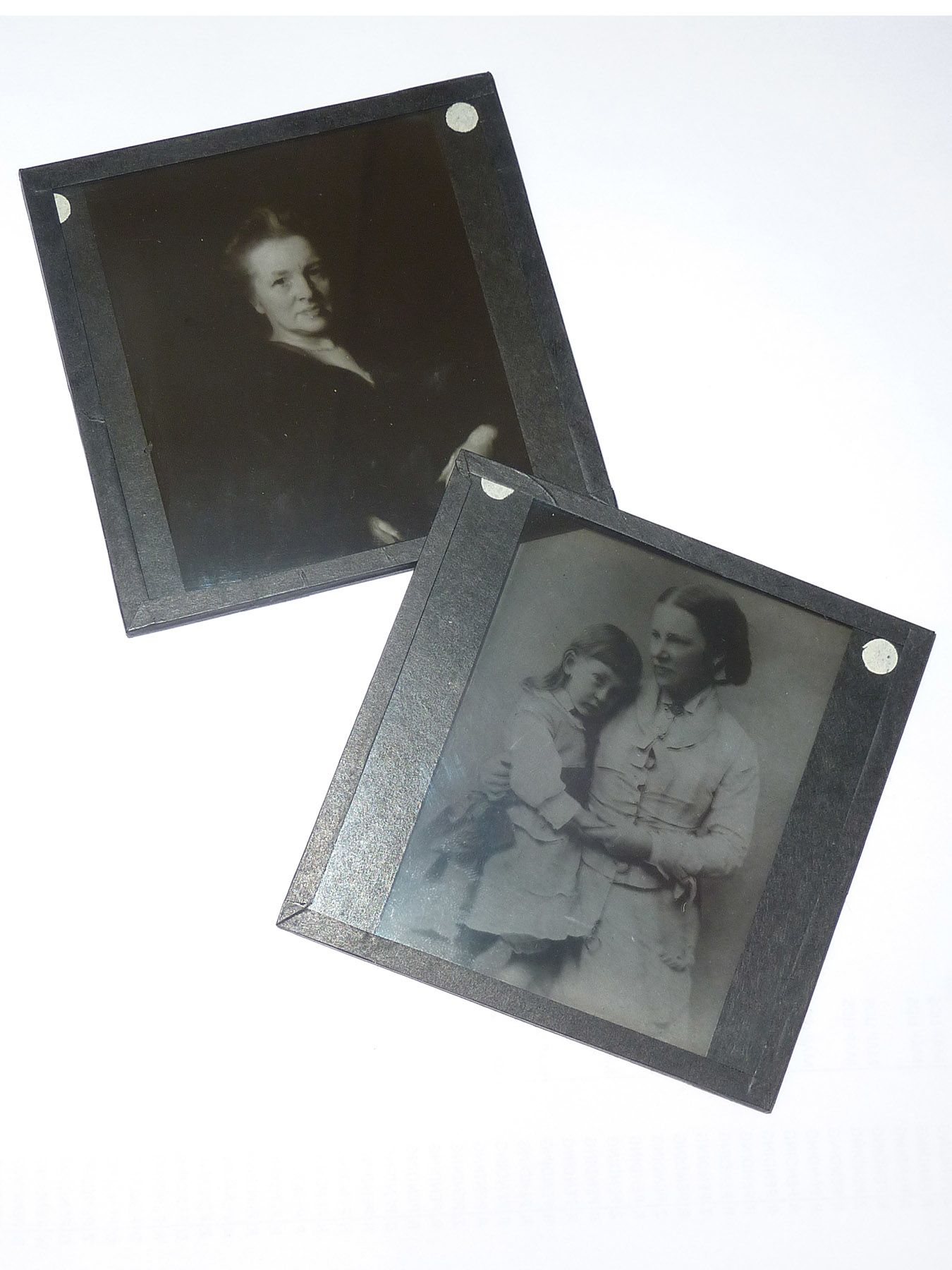 Photograph of two glass lantern slides. One shows a portrait of an older woman in a dark formal dress. The other is a portrait of a woman holding a child on her knee, both dressed in light coloured clothing.