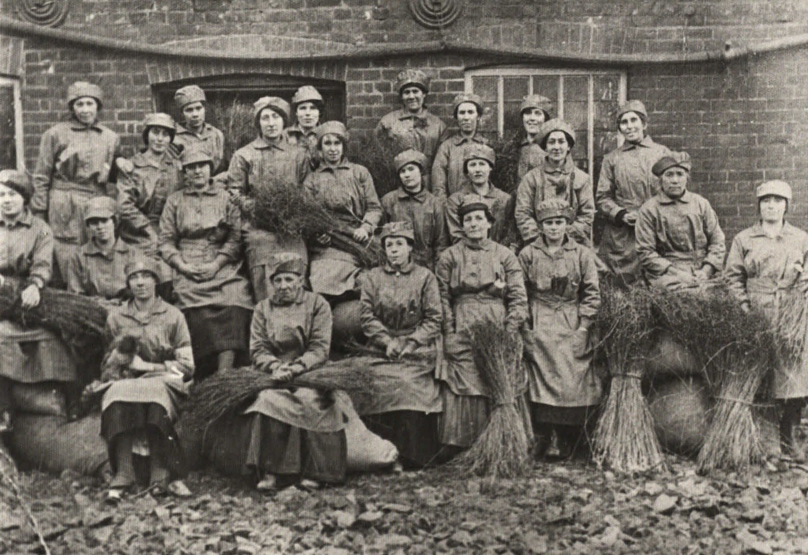 Black and white photograph of a group of women arranged in three rows in front of a brick building. The women are wearing work tunics and cloth hats, and several of them are holding bundles of flax.