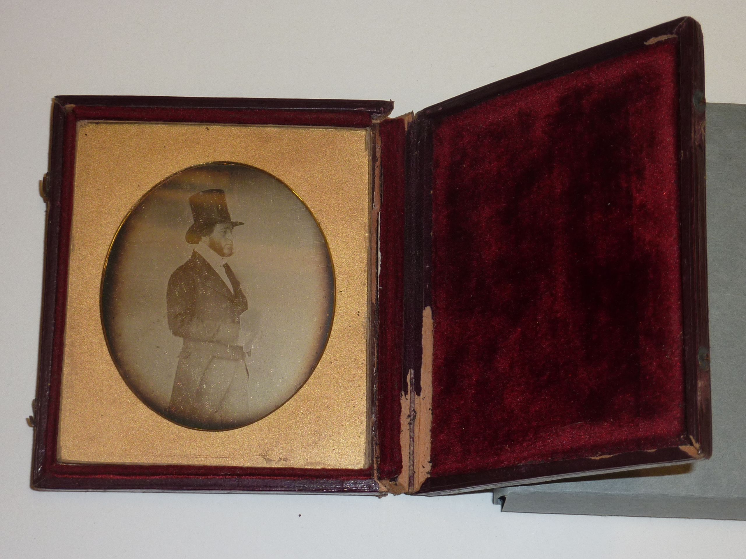 A case which opens like a book which contains a sepia photograph of a man in formal clothing, including a top had and tails. He also has very long and fluffy sideburns. The image is surrounded by a golden frame with an oval aperture. The other side of the case is lined with red velvet.