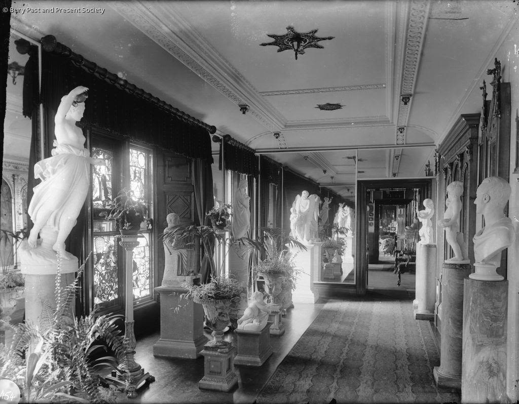 A greyscale image of statues filling the entire corridor of the house. No space escaped decoration. Windows, with sunlight blasting through them are also present. 