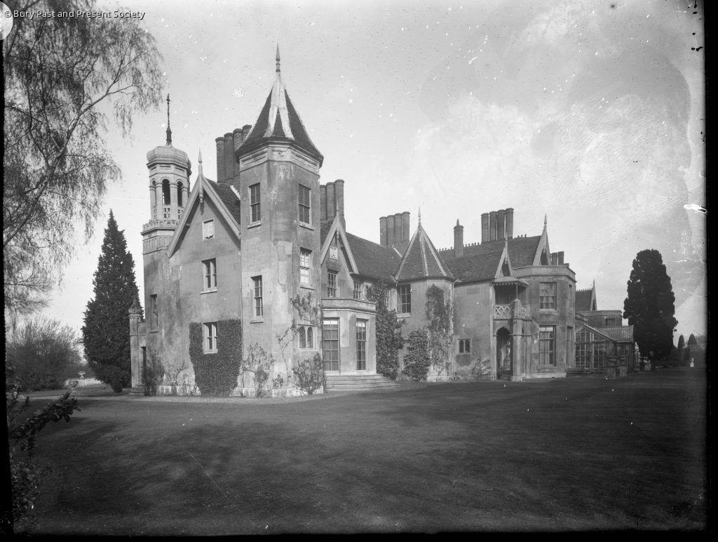 A black and white photograph showing the rear, western end of Hardwick House. There are a variety of roof shapes, and multitudes of windows, chimneys and entrances. Grass lawn, and occasional trees are also present. 