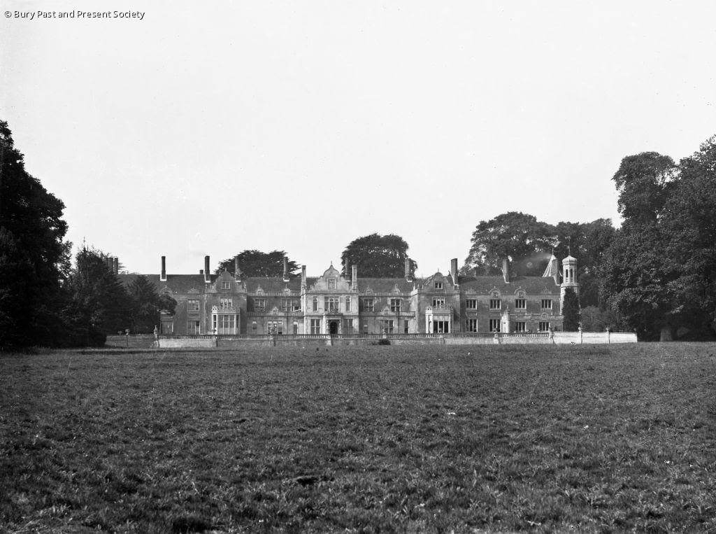 A black and white image of Hardwick House taken from the front with multiple chimneys, windows, and entrances. A large grass field and trees, fully leaved are also present. 