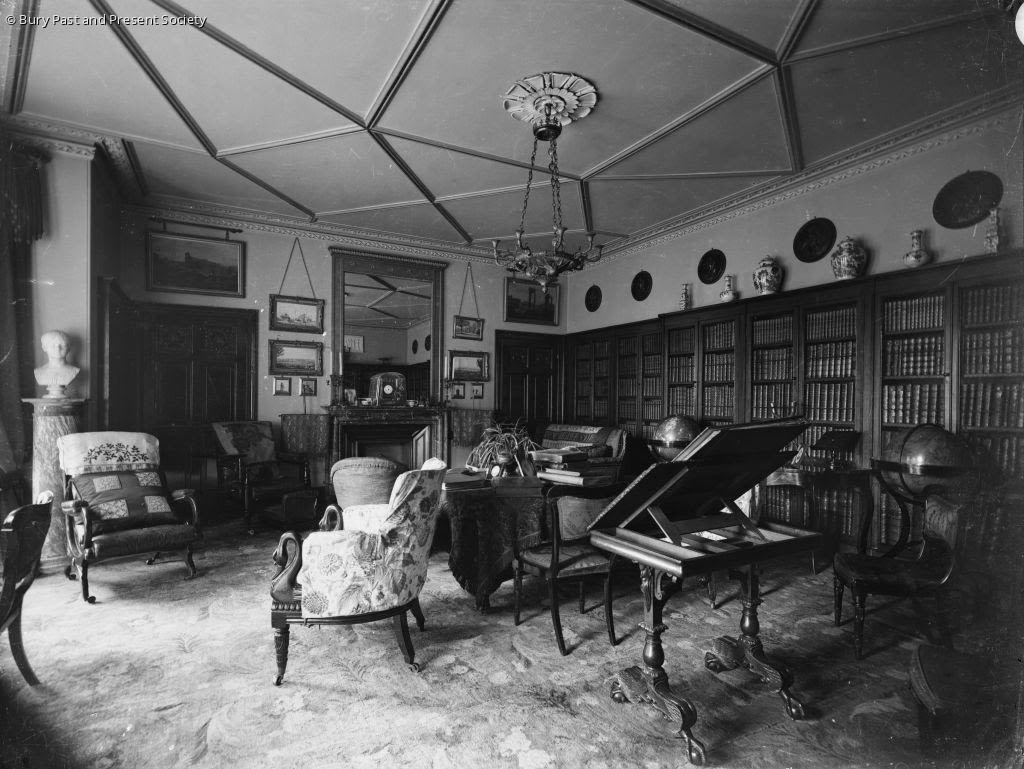 A greyscale image of the interior of the library room in Hardwick House. No surface escaped decoration with chairs, tables, a piano, bookcases, and pictures adorning the walls.  