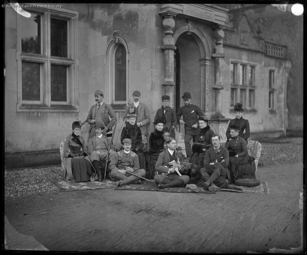 A greyscale image showing members of the Cullum family gathered round the pouch of the house. Both women and men, including George Gery Milner Gibson Cullum who is seated centre with a dog are present.  