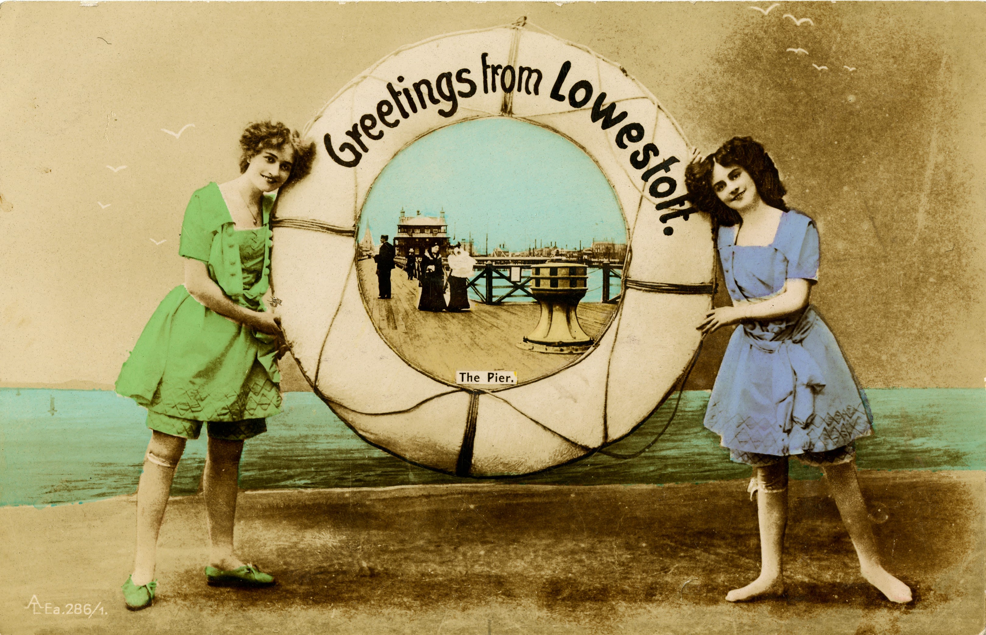 A full colour postcard with two ladies, one on the left wearing a green dress and green shorts and light brown tights and the other one, on the right, wearing a blue dress and shorts and light brown tights. They are both standing as close to as possible a lifesaving rubber ring which has the following text at the top “Greetings from Lowestoft”. Inside the ring is a view of south pier, Lowestoft with the words “The Pier”. The pier has a brown wooden decking, fence, and main pier house visible. To the left of the pier is the main esplanade visible. There are two people on the pier which are standing together, in conversation. One is wearing white top and black trousers and the other is wearing an all-black dress. Behind them is a man looking out who is wearing a black suit and behind that is another person who looks to be wearing an all-black outfit. 