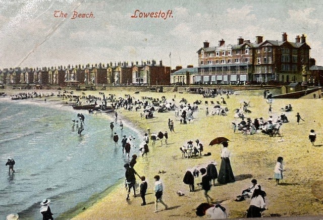 A full colour postcard of the esplanade and beach of Lowestoft. In the foreground is the sea, with the yellow-coloured sand further back.  The beach has hundreds of people on it, all wearing different outfits and all doing different activities. Some of them are playing in the sea, others are chatting, others are children talking to parents and others are sitting on the beach. Some even have their deck chairs out. Behind the people are a row of houses, with all of them having brown brickwork and multiple chimneys. Separating the beach and houses is a sea wall which stretches into the distance. 