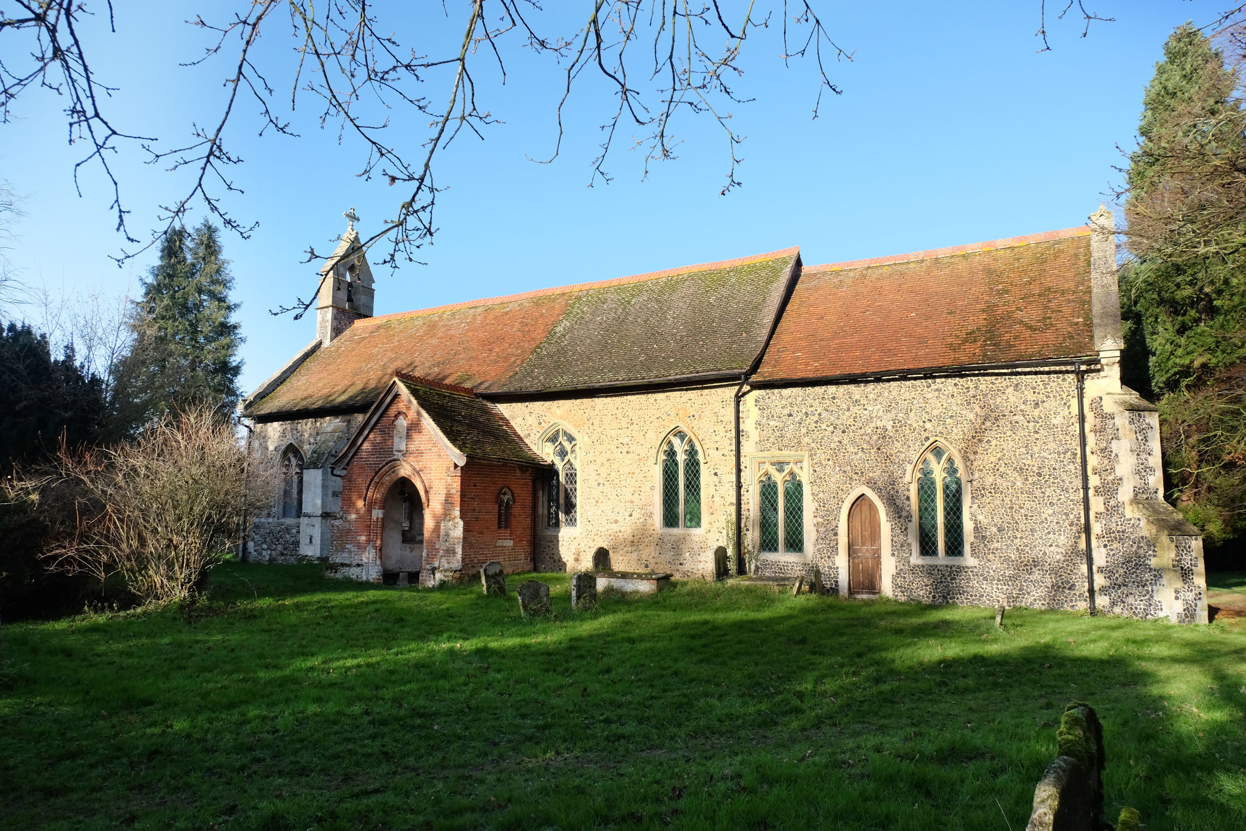 Colour photograph of the outside of parish church showing porch and priests door into the chancel. There are trees and shrubs at either end of the church.