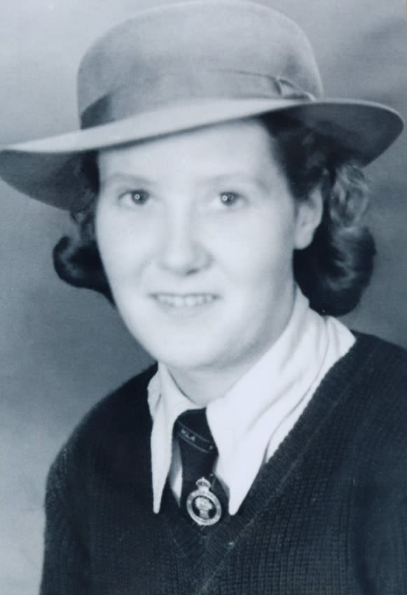 Black and white portrait photograph of Grace Eveline Launt. Her head and shoulders are in the photograph, and she is looking at the camera and smiling. She wears a v-neck jumper over a shirt and tie, which has the Women's Land Army badge pinned to it. Her hair is curled and goes just past her ears, and she is wearing a hat.