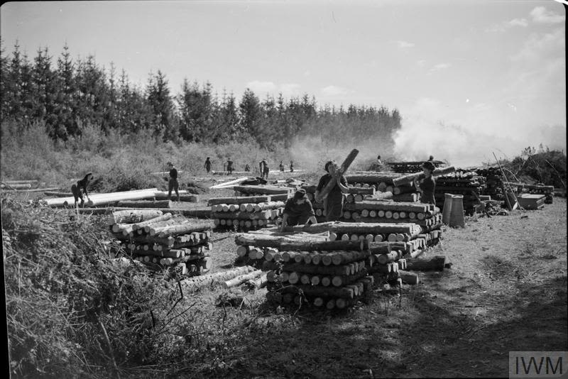 Black and white long shot photograph of several land girls stacking pit props. There are around 8 visible stacks, which have several props within them. There is smoke towards the background of the photograph
