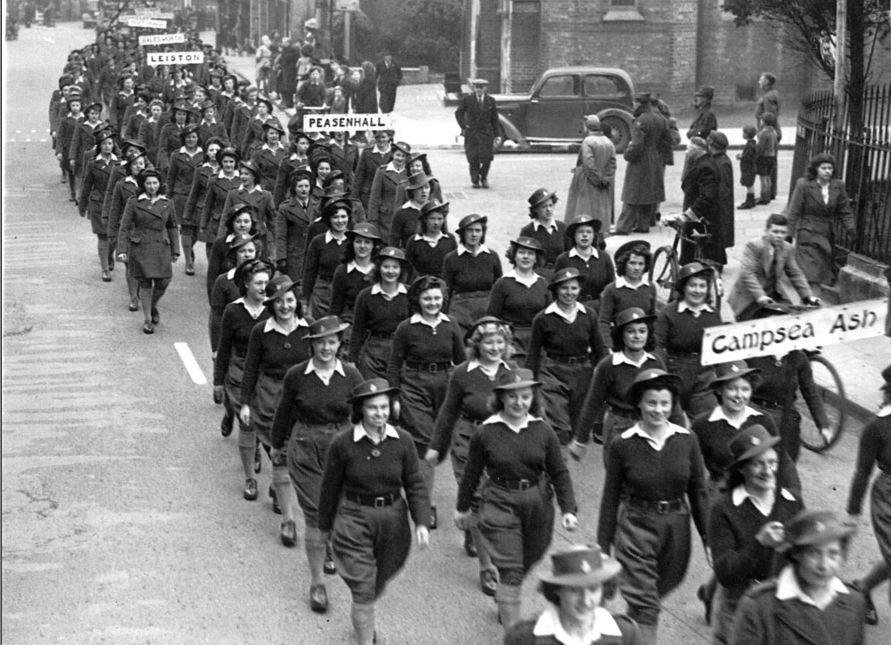 Black and white photograph showing four columns of women in Land Army uniform marching through a street. They hold signs to show where they are from. In the foreground a sign says Campsea Ash, and in the background is one for Peasenhall. The women are smiling and some of them are looking at the camera.