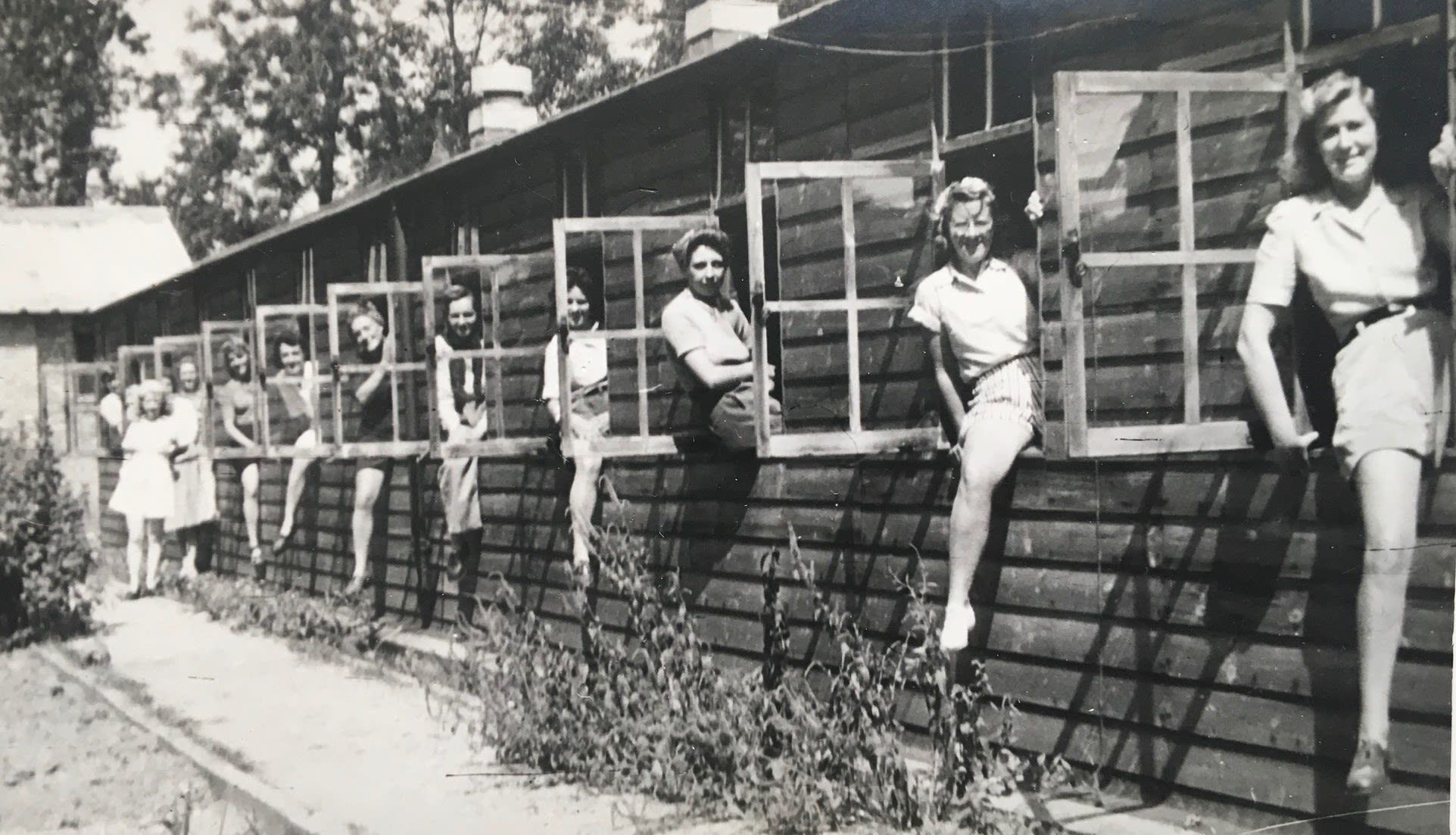 Black and white photograph of a long, low wooden hut with several windows. All of the windows are open, and a young woman is sat in each window with one leg inside the building and one leg out, all looking at the camera and smiling.