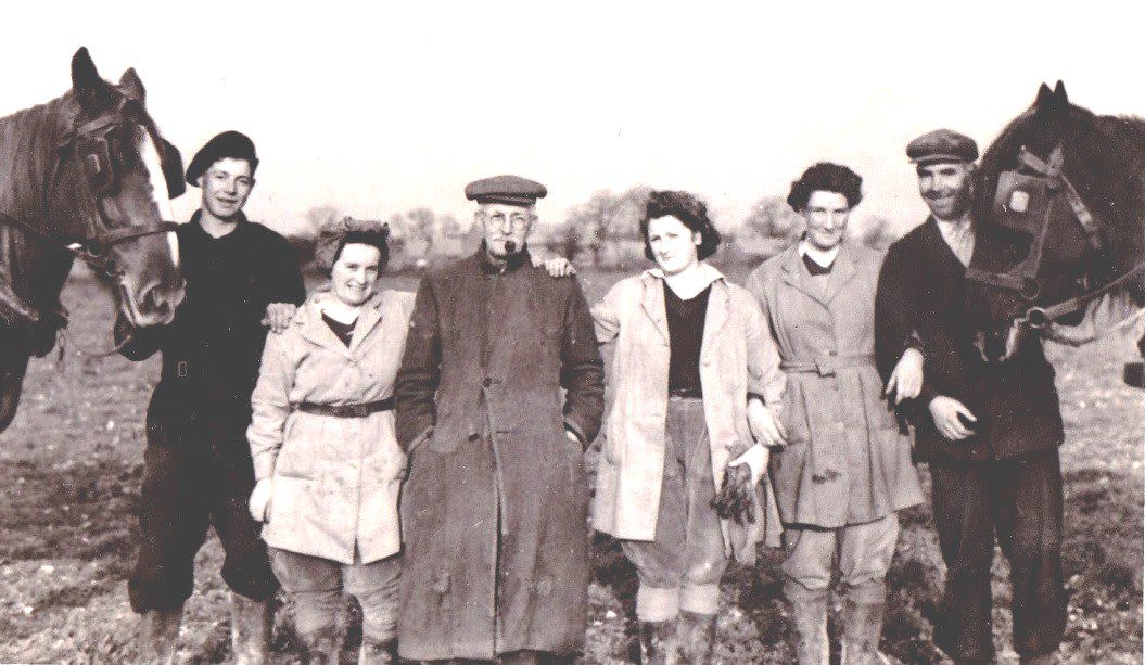 Black and white photograph of Vivian, alongside 3 male farm workers and two other Land Girls. There are also two horses. The six people stand in a line and all face the camera and smile, with the two horses either side of them. They are all wearing winter jackets and breeches. All three men are wearing berets and the man in the middle is smoking a pipe.