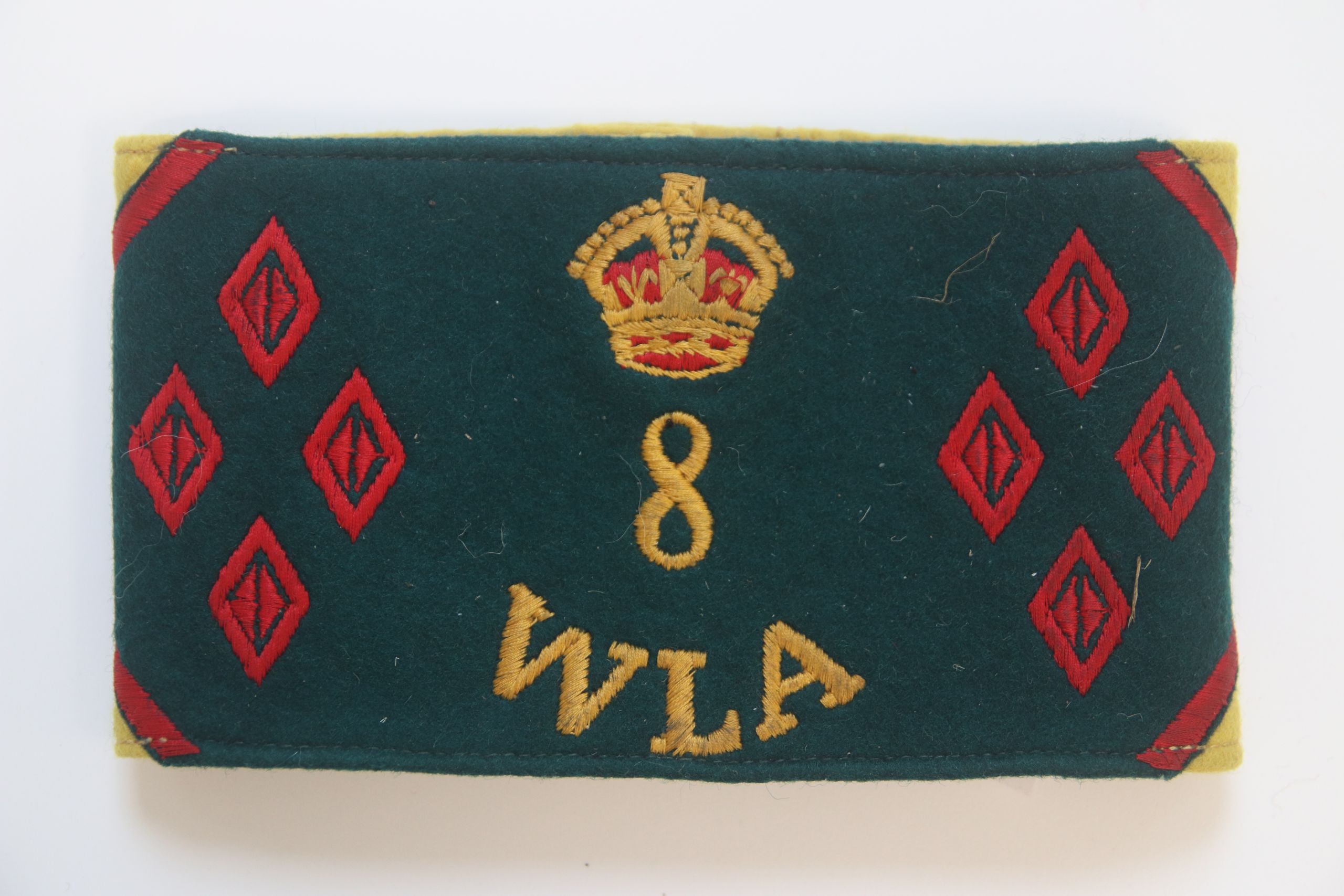 Colour photograph of a Women's Land Army armlet. It has a dark green background. In the centre is a gold and red crown. Underneath the crown is a golden number 8 and the initials WLA. On both the right and left sides are four red diamonds.