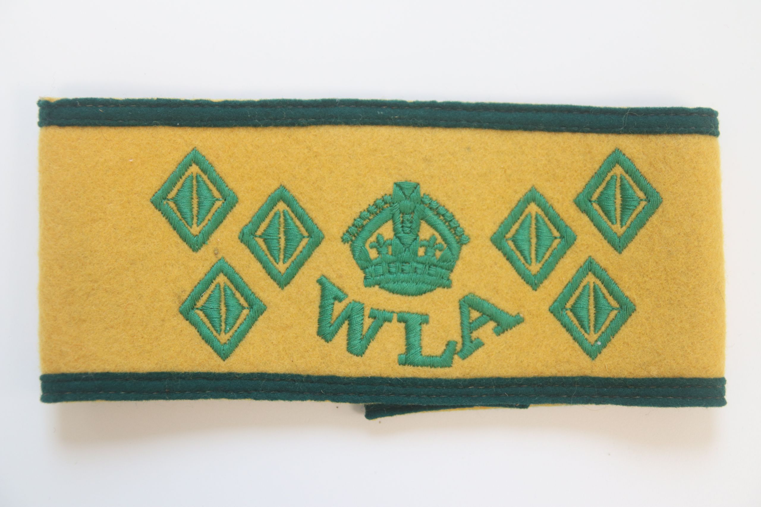 Colour photograph of a Women's Land Army armlet. It has a yellow background and dark green edges at the top and bottom. In the genre is a green crown, and below it the initials WLA. On either side are three green diamonds (six in total).