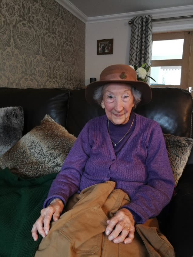 Coloured photograph of Audrey Baxter in March 2020. She is sitting on a black leather sofa with her hands on her lap and is looking at the camera and smiling. she is wearing a purple top and cardigan, and has grey hair. On her head is a brown felt Land Army hat.
