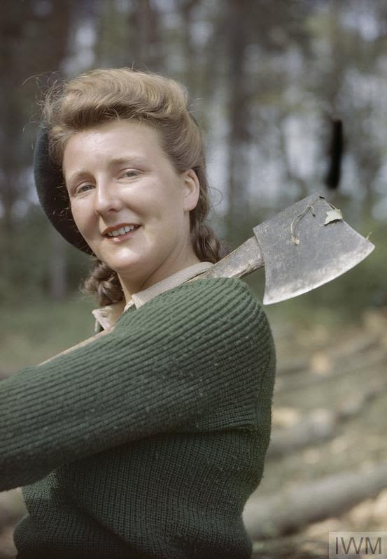 A coloured photograph of Vera Howson, a Lumber jill. She is close to the camera, with only her top half in the photo. She is wearing a Khaki coloured jumper and a beret on her head. She is holding an axe over her shoulder.