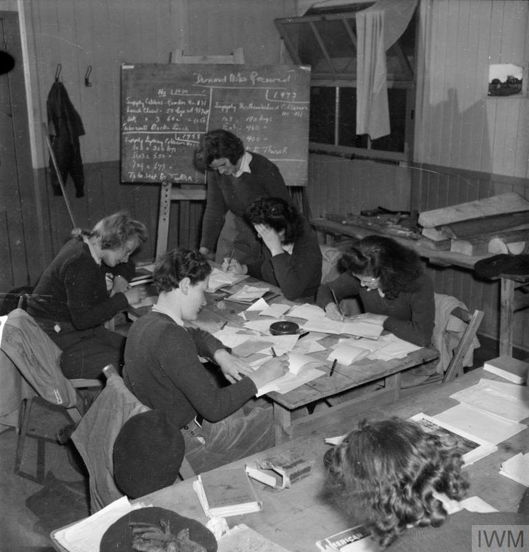 A black and white photograph of 5 land girls sitting an exam as part of their training. 4 of the girls sit at one desk, and are looking down at their papers, while another woman stands over the desk and watches them. There is a black board behind them with some writing on it, as well as another desk with some books scattered across it.