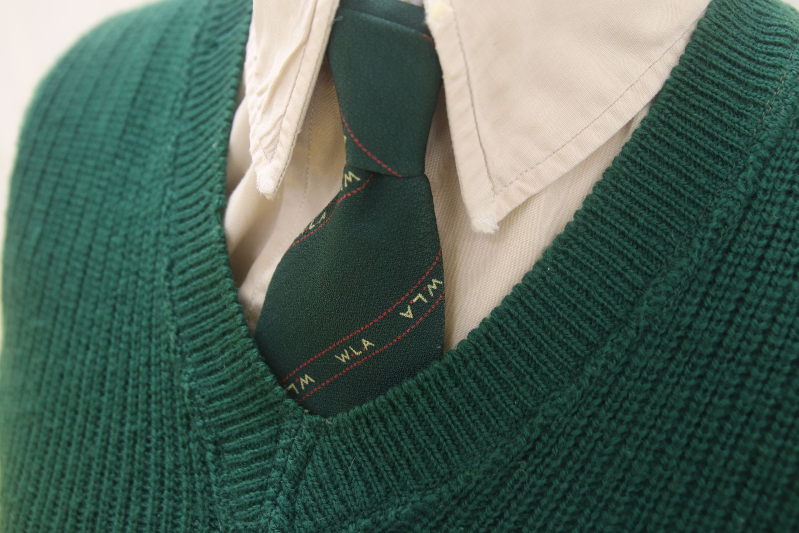 Colour photograph showing a close up of the green V-neck Women's Land Army jumper, worn over a cream cotton shirt and Women's Land Army tie. The tie has a dark green background, with diagonal stripes across it. The stripes are made up of very thin red lines, with the initials WLA in between them in yellow.