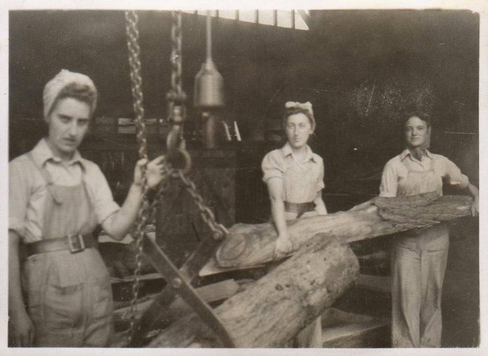 Black and white photograph of three women working in a timber mill. They are moving a large, long log using chain hoists. They are all wearing the Land Army polo shirts and dungarees and have their hair tied in scarves.