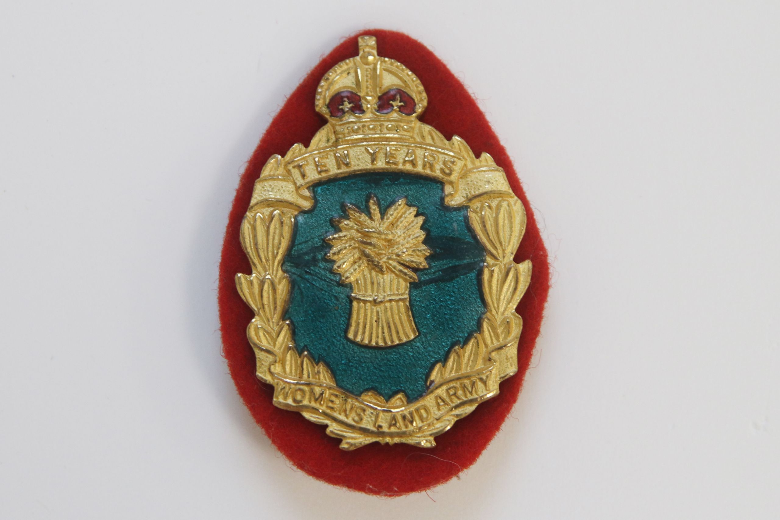Colour photograph of an enamel badge. The main part of the badge is oval in shape. On top of the oval is a gold and red crown. The outside of the oval is gold, and contains the words Ten Years and Women's Land Army. Inside this golden band is a dark green enamel, containing a golden sheaf of wheat.