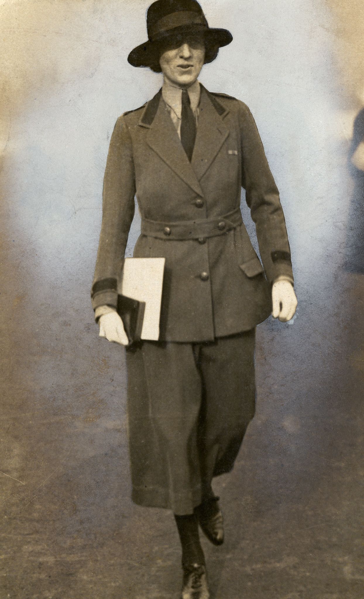 Sepia photograph of a women in a smart suit with a long skirt, walking towards the camera. She is wearing a dark felt hat, and carrying a book and bundle of papers in her right hand.