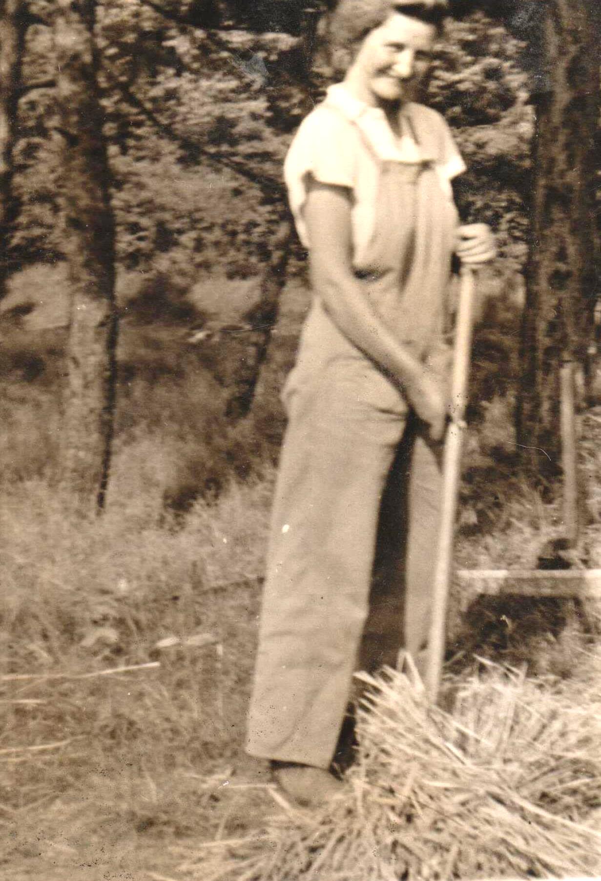 Sepia photograph of Vivian on her 21st birthday. She is wearing dungarees over a white short-sleeved top, and is looking at the camera and smiling. She is holding a wooden stick, which could possibly be a rake, and at the bottom of it is a pile of straw which she has gathered.