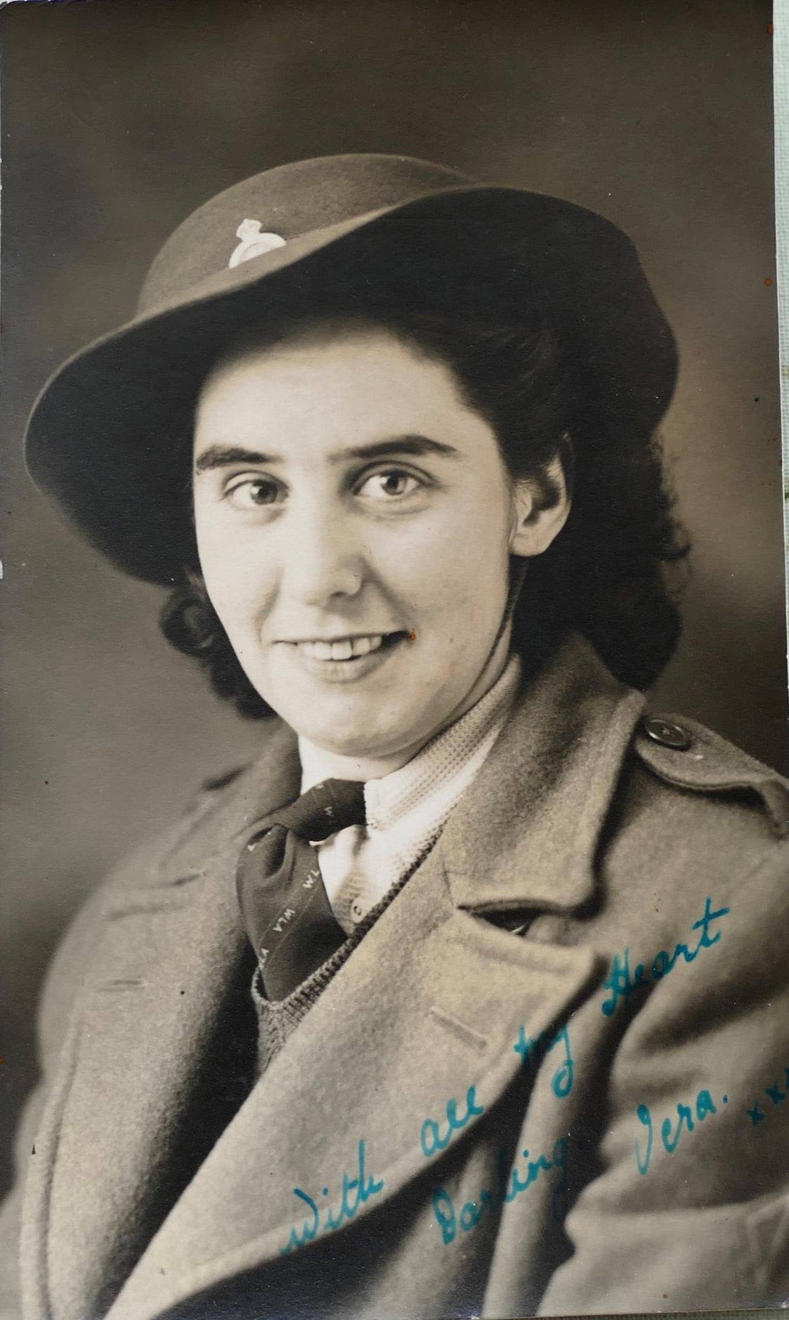 Black and white head shot of a young woman in Land Army uniform, including her thick great coat and hat. Her curly dark hair spills out from under the hat