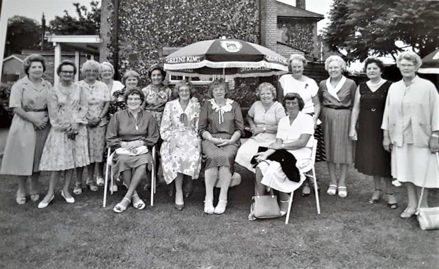 A black and white photo of 15 ladies sitting in a pub garden under a sun umbrella. They are smartly dressed and smiling at the camera. From their clothes the picture looks to date from the 1970s.