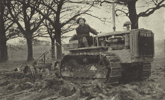 Sepia photograph of a Land Girl steering a Caterpillar tractor and looking in front of her. She is in a field and there are some trees in the background. She is pulling a cart behind the tractor.