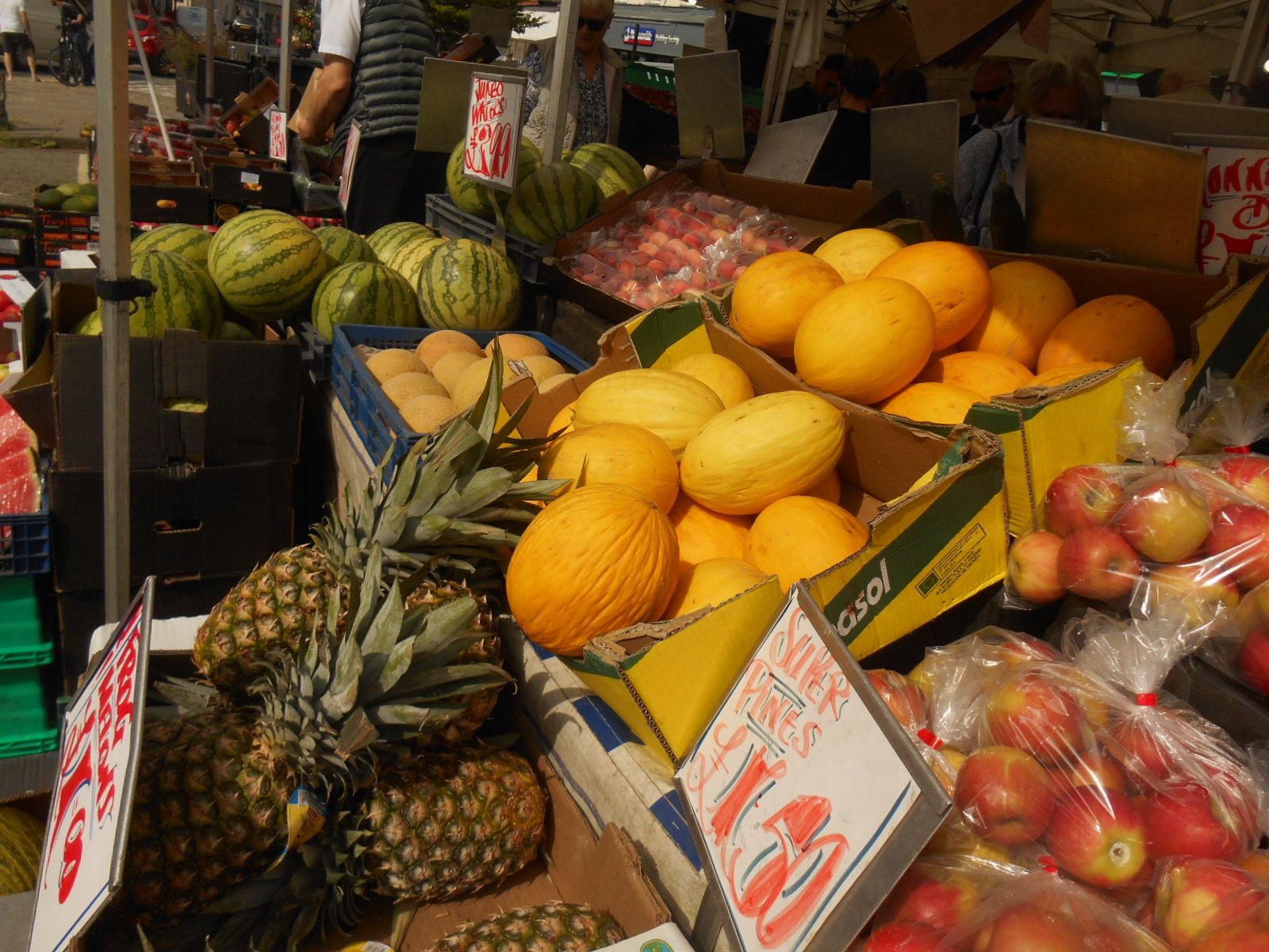 A colour photograph of bright sun shining on a fruit stall. Particularly prominent are golden-coloured melons, and beneath them the spiky crowns of pineapples.