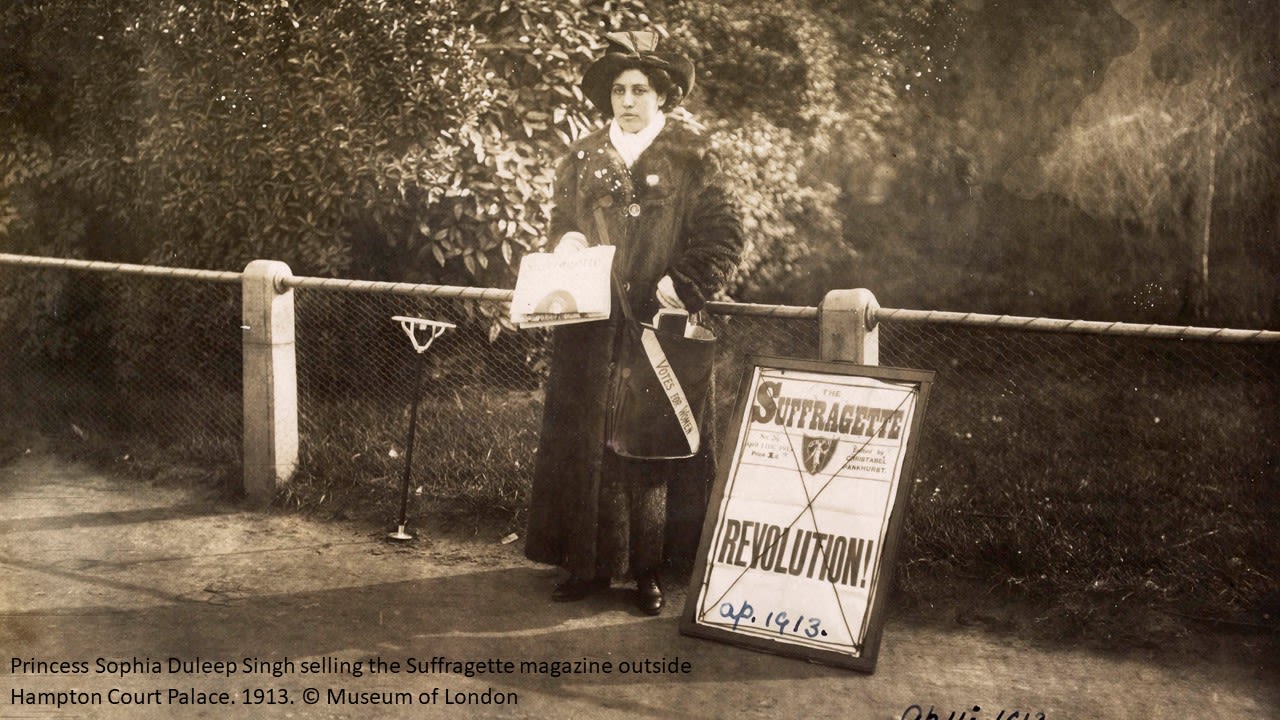 An image of Princess Sophia Duleep Singh who is standing outside Hampton Court Palace in 1913 trying to sell the Suffragette magazine. She has a billboard to the right of her advertising the magazine. 