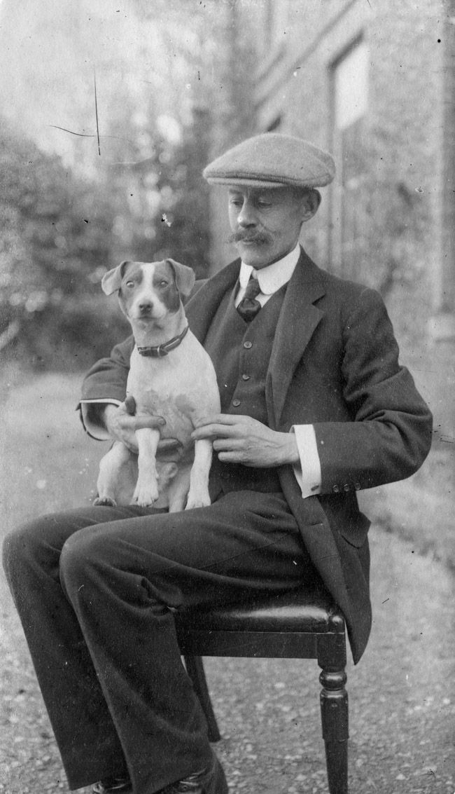 Black and white photograph of a man in a suit and flat cap sat on a chair in front of a large house, with a small dog on his knee