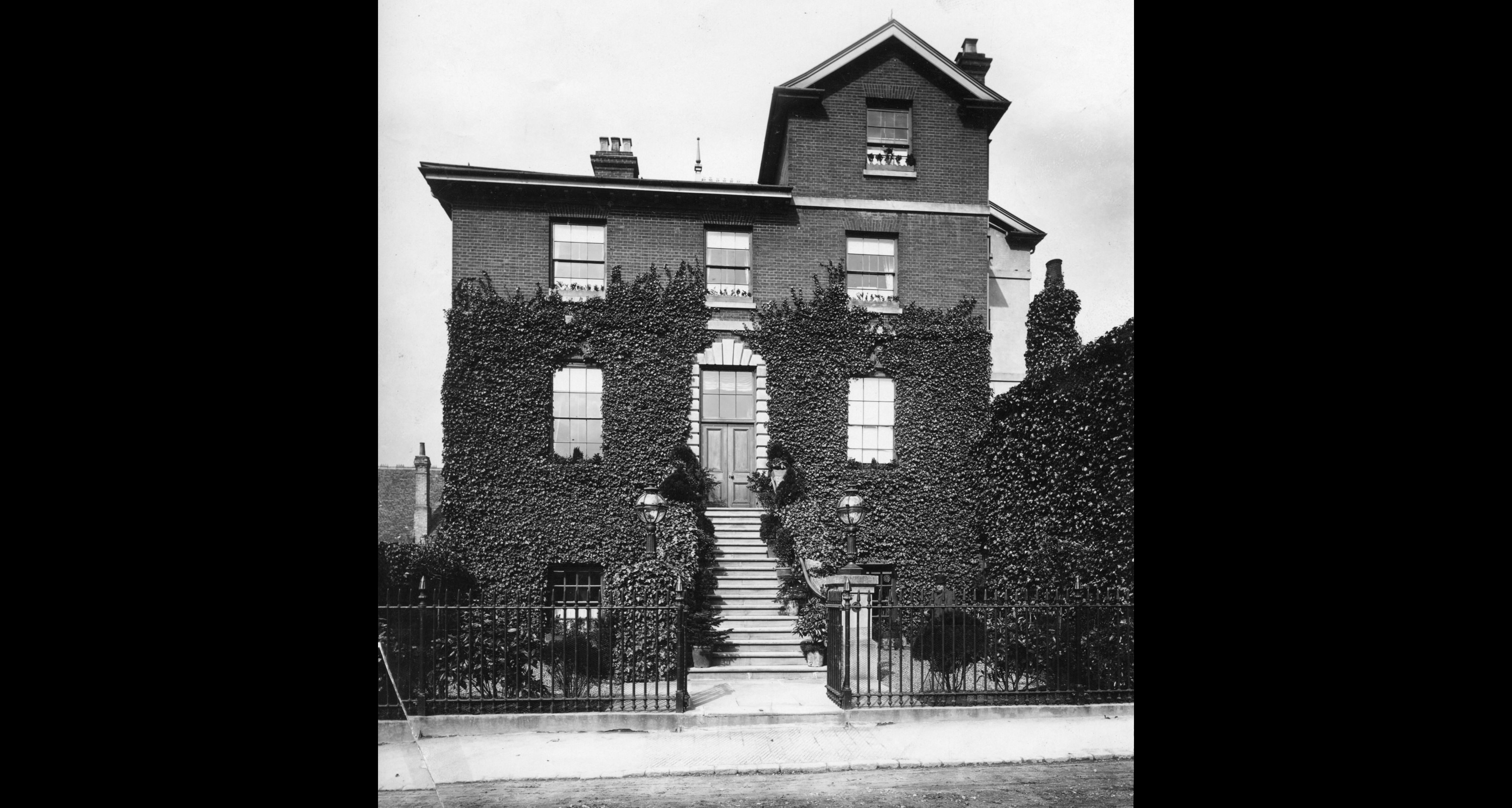 Historic black and white photograph of a large, very smart brick house of three storeys. It has a large dormer in the roof and a flight of stone steps leading to a large entrance door on the first floor.