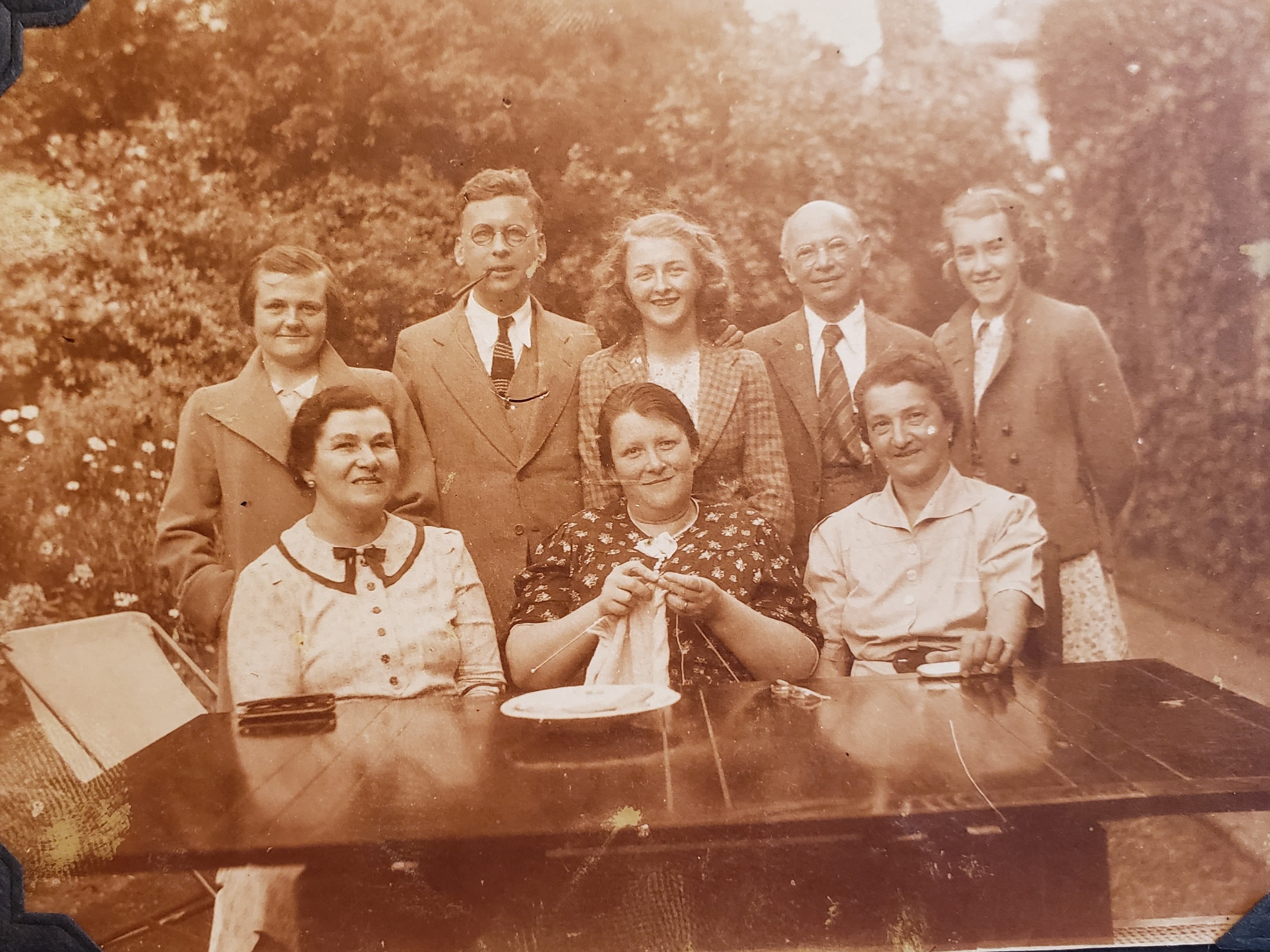 Sepia photograph of a group of 8 people smiling at the camera. 5 people stand behind 3 people seated at a table in a garden. There are 6 women and 2 men. One of the men has a pipe in his mouth, and one of the women is knitting. They are all dressed in 1940s clothing.