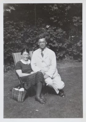 Black and white photograph of a man and a woman on a deck chair in a sunny garden. The chair is on a lawn and behind them is a leafy flower bed. The woman is sitting on the deck chair and the man is perched next to her. She has a knitting bag by her side.