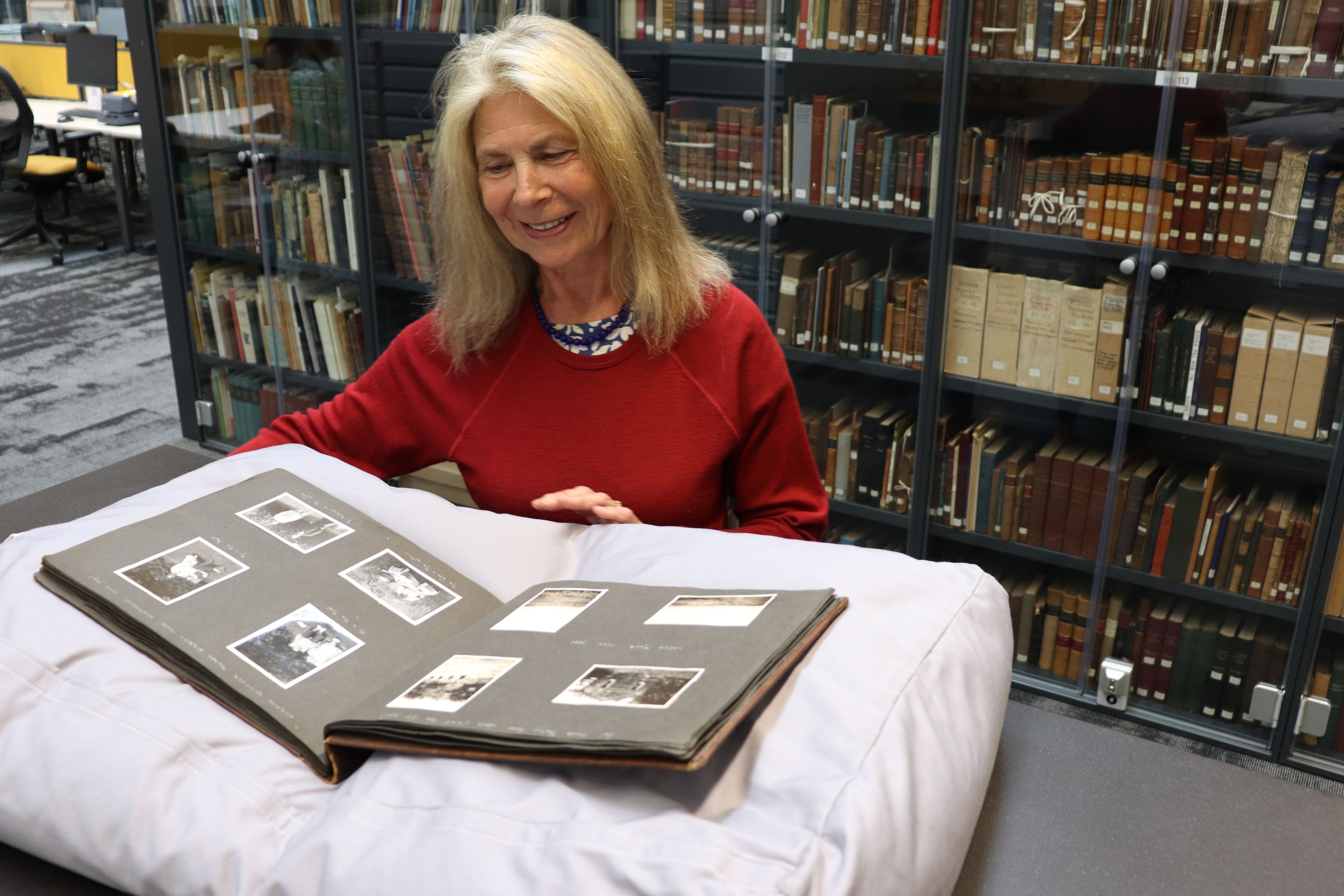 Modern colour photograph of a woman with blonde hair in a red jumper stood in front of shelves containing historic books which are safely behind glass doors. She is looking at an old photo album in front of her, which is supported on a large grey cushion.