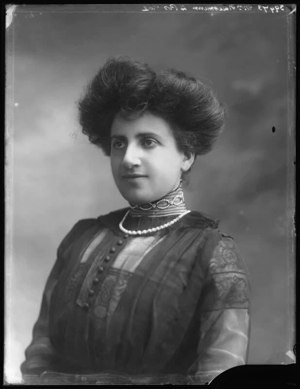 A sharp black and white historical portrait photograph of a young woman in luxurious, smart Edwardian clothing. Her dress includes a sheer overlay and a very high neck that comes to under her chin. Around her neck is a string of pearls. She has fair skin and dark hair which is styled in a large crown about her head.