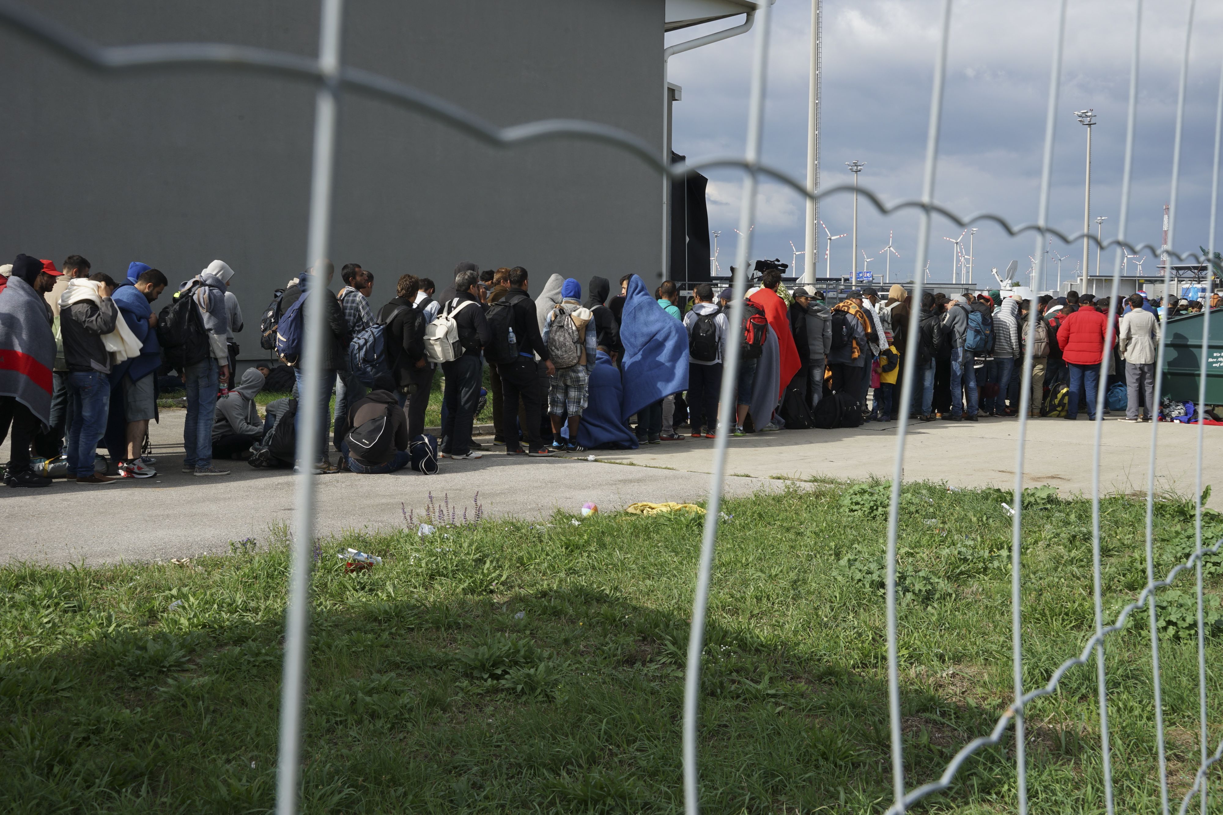 Modern colour photograph of people queueing behind a wire fence. There is a large number of people, many with bags or suitcases. They are wearing warm clothes and some are wrapped in blankets.