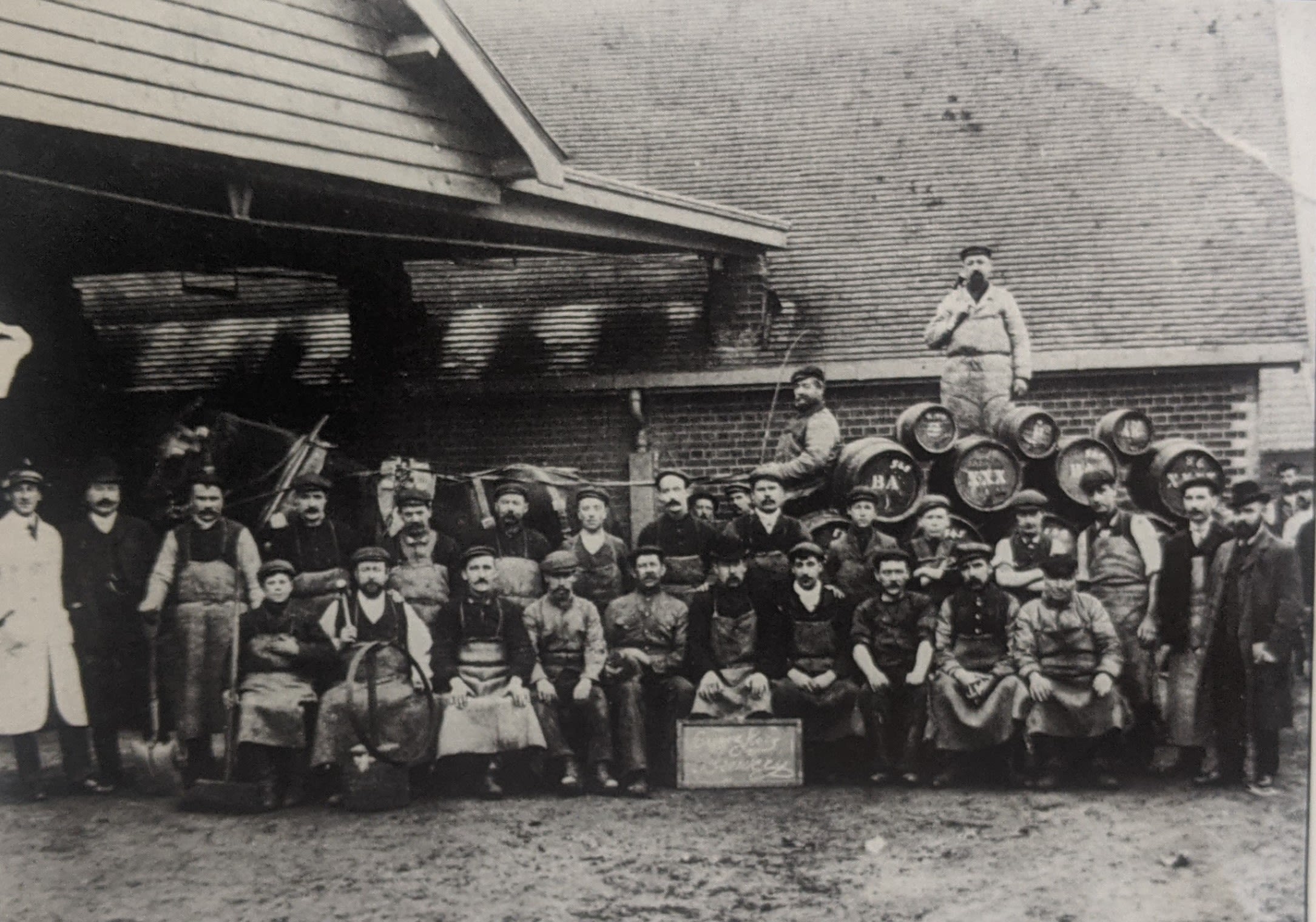 Black and white outdoor photo of a group of men and boys posing around a horse-drawn cart loaded with barrels