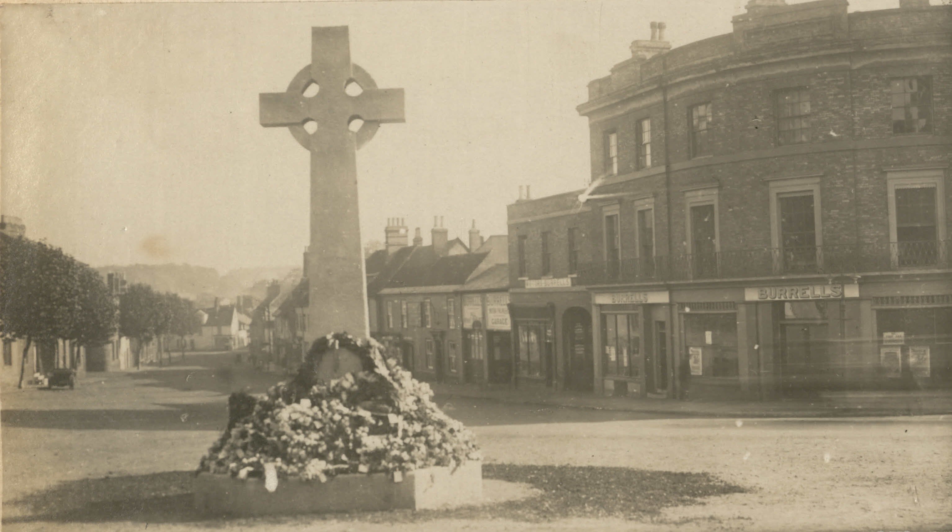 Sepia photo of a view down a street of historic buildings, with a large stone cross in the foreground. Its base is piled with wreaths of flowers.